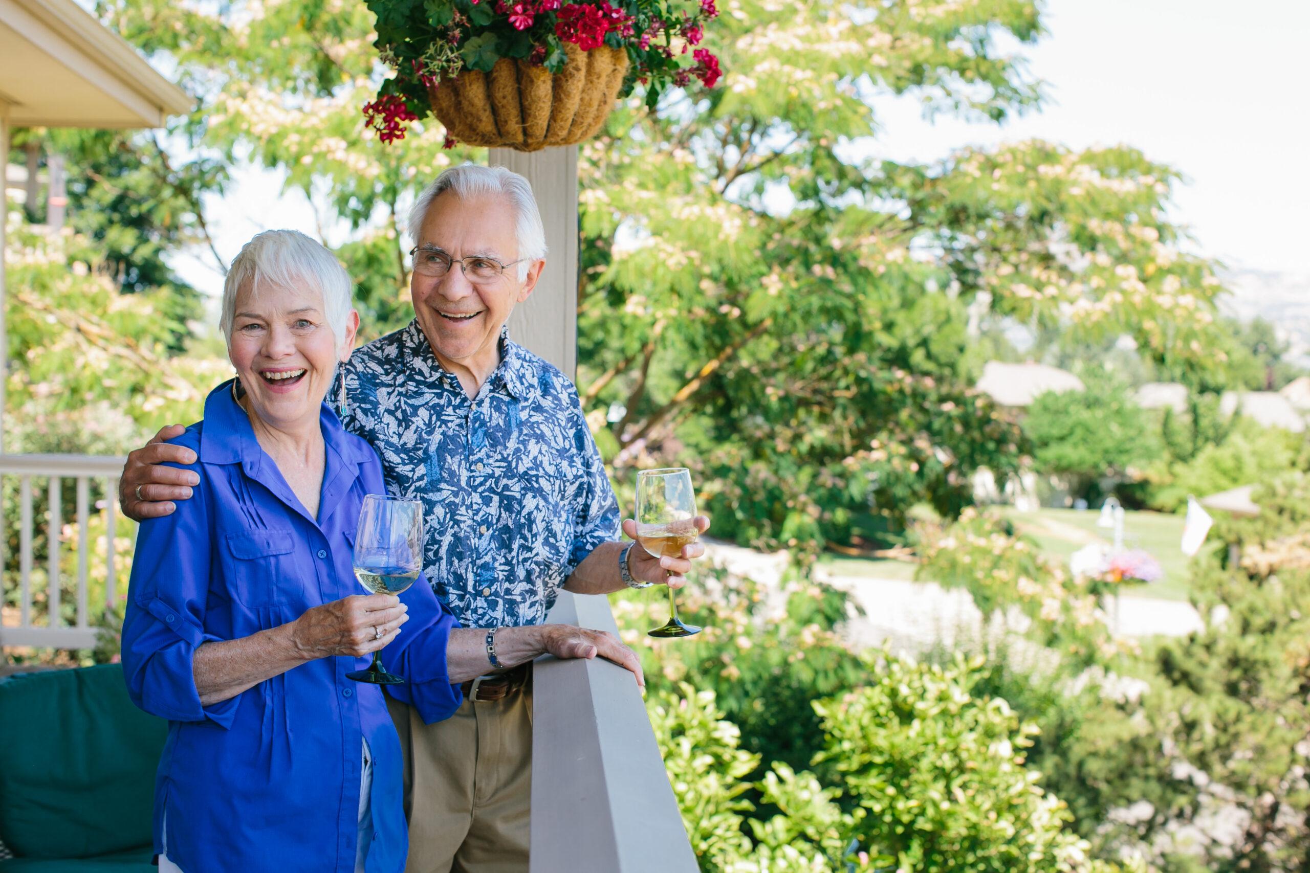 An older couple holding wine glasses smiles on an outdoor patio.