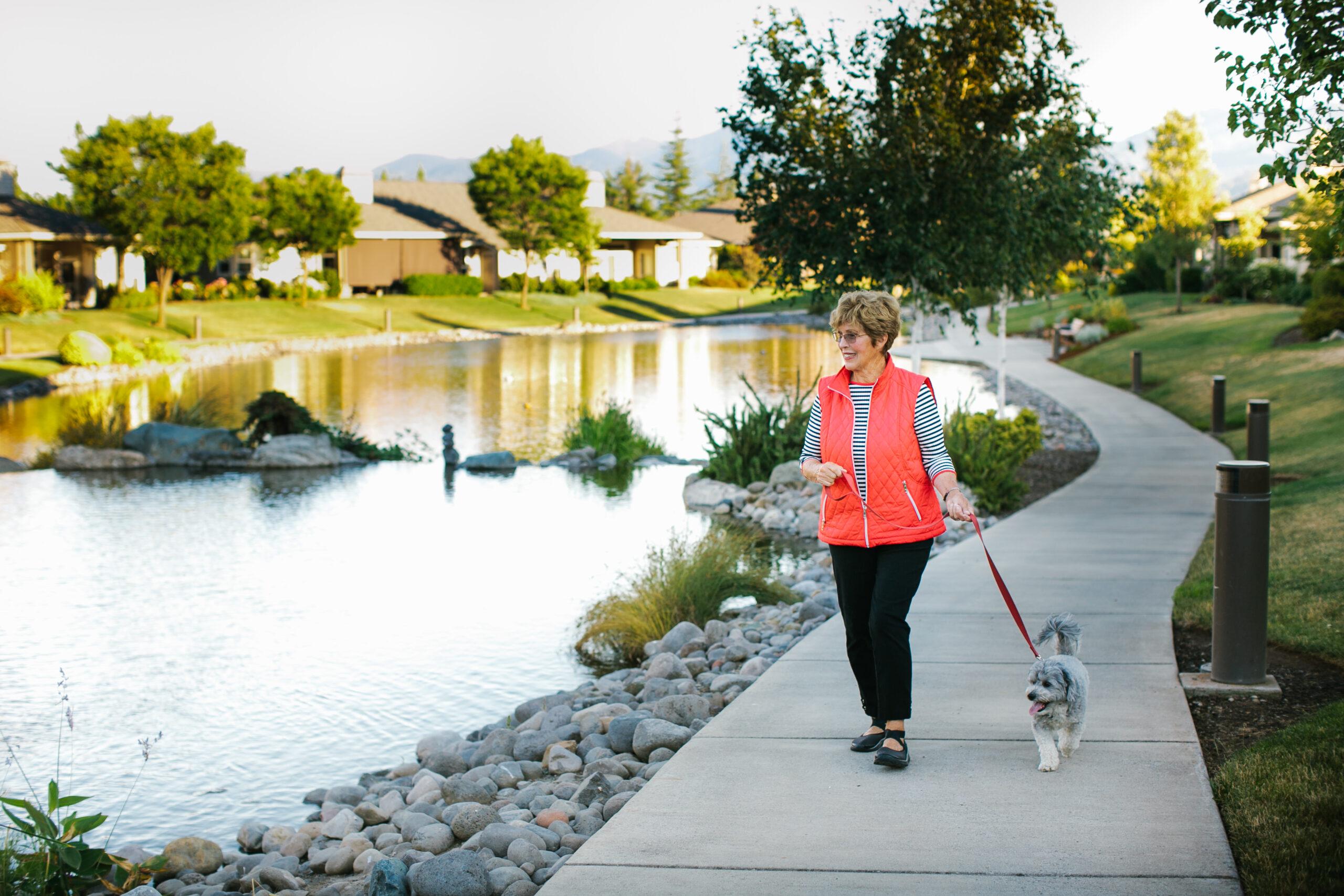 A woman wearing a coral colored vest walks a dog next to a picturesque lake.