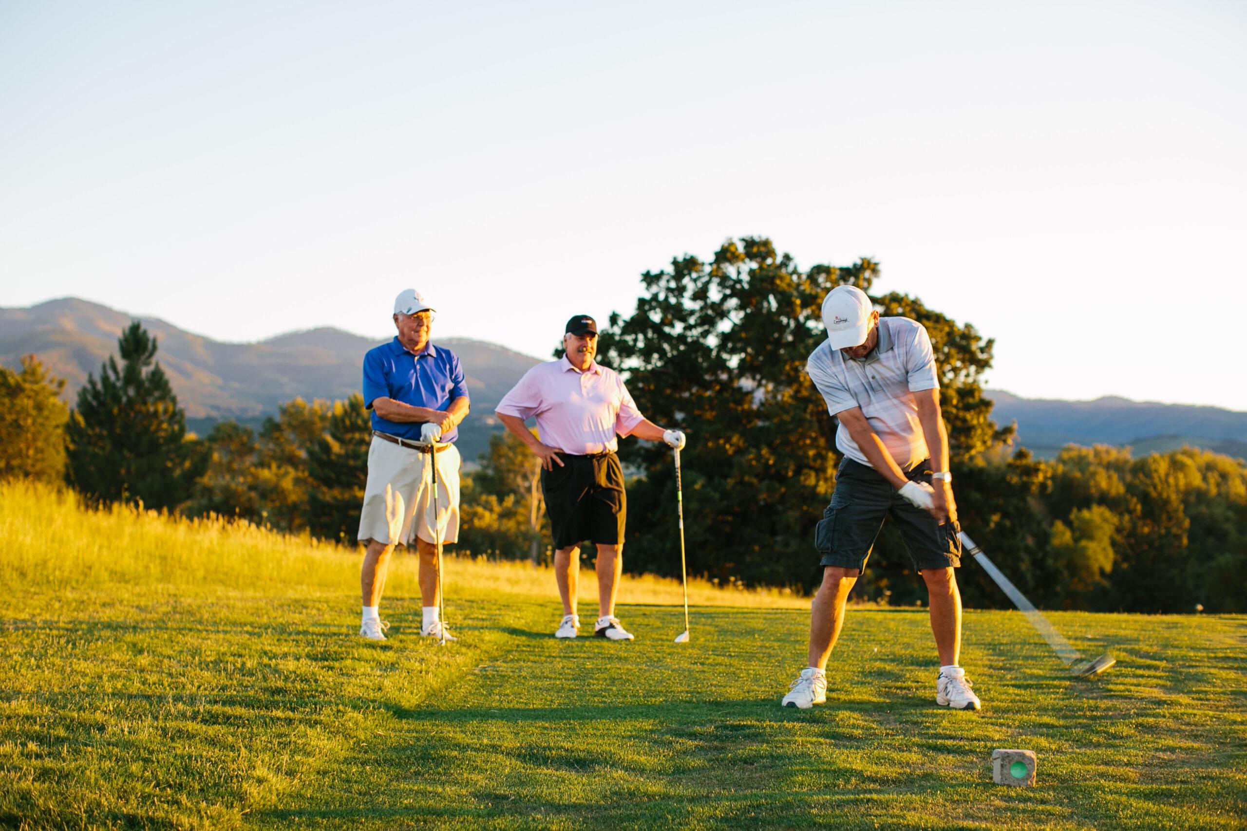 A man swings a golf club as two nearby men watch during golden hour.