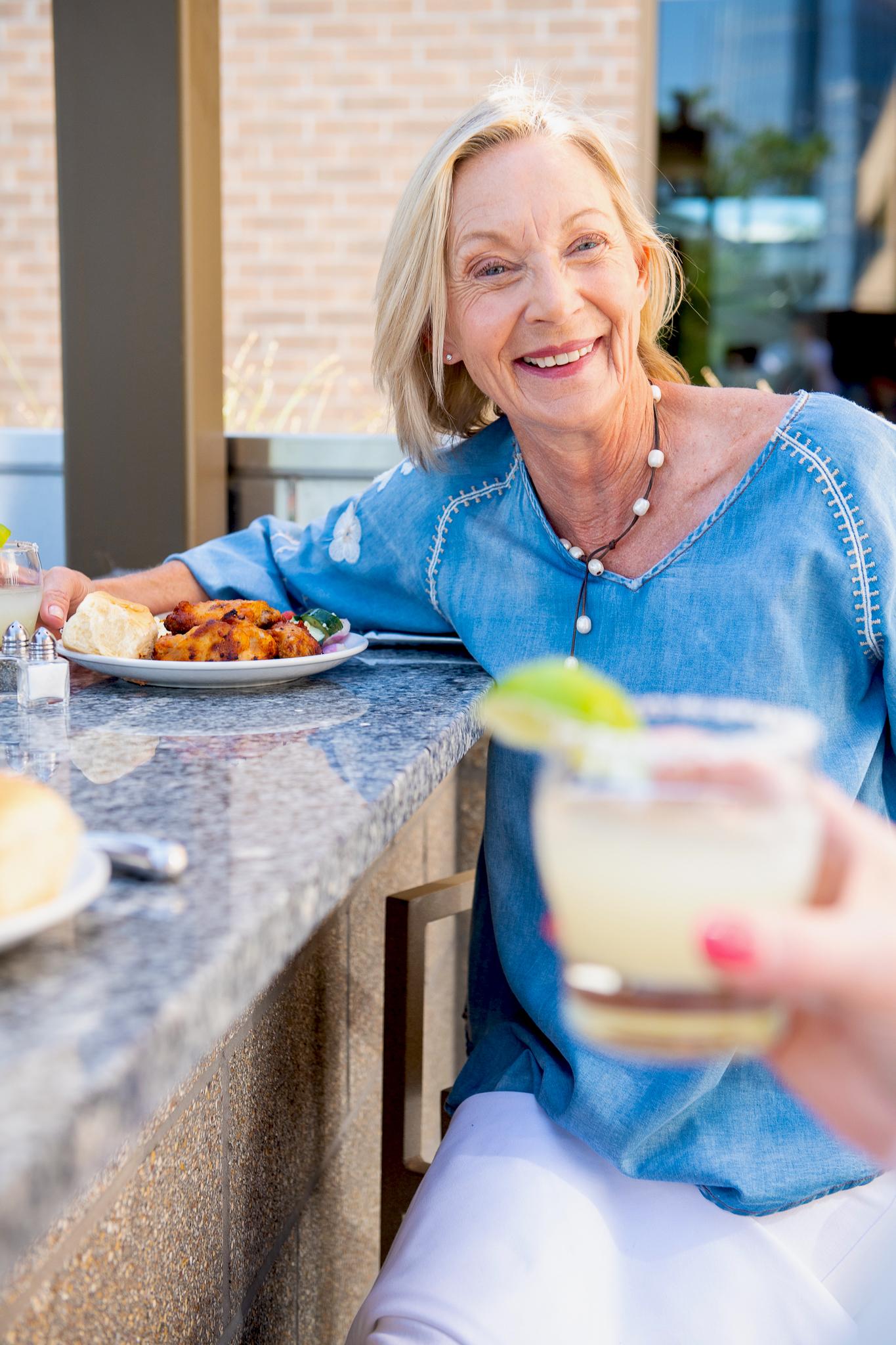 A happy woman eats a meal outdoors. Someone is holding a drink with a lime and salted rim in the foreground of the photo.