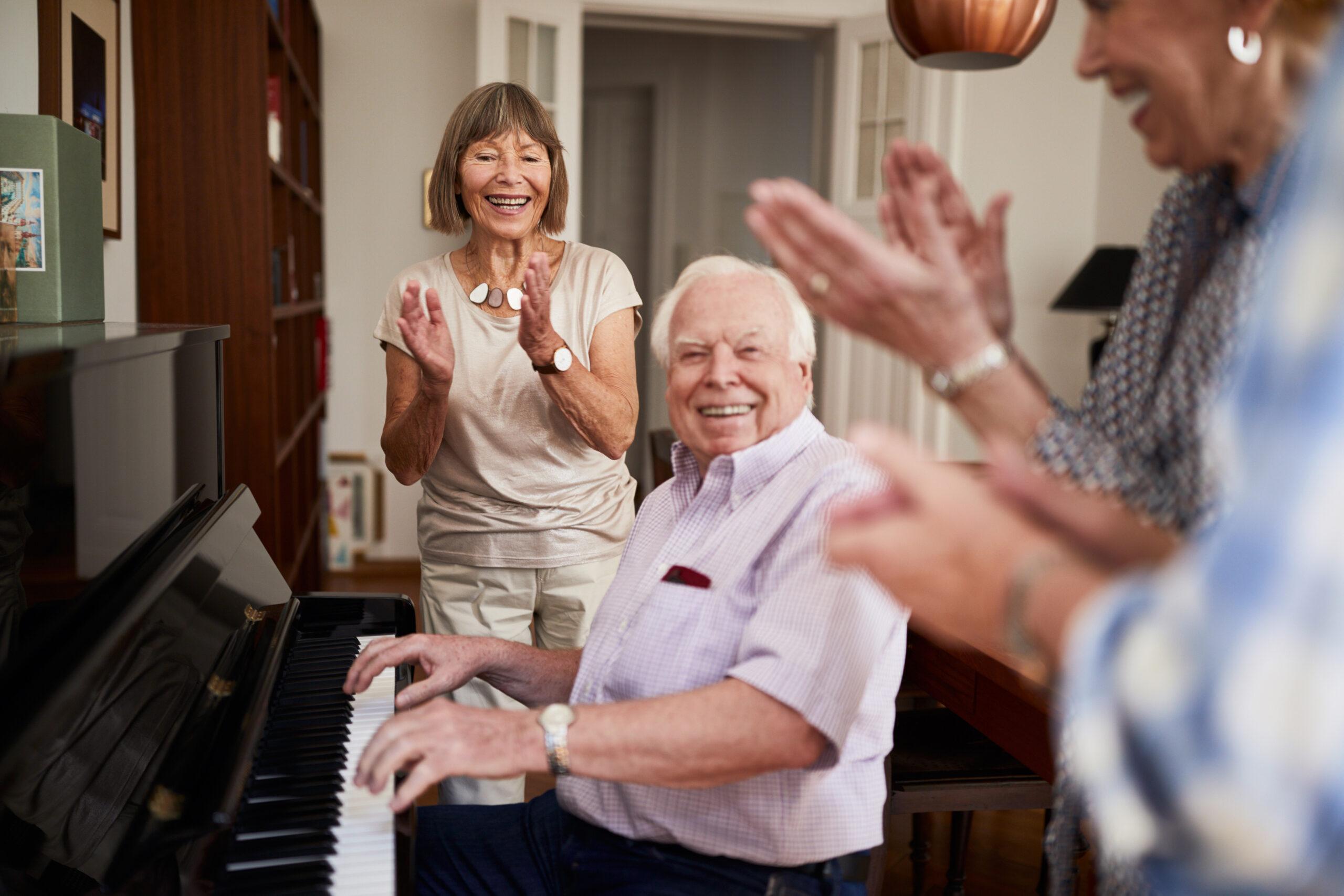 Happy seniors enjoying a small get-together. A man plays piano while his friends clap to the rhythm.