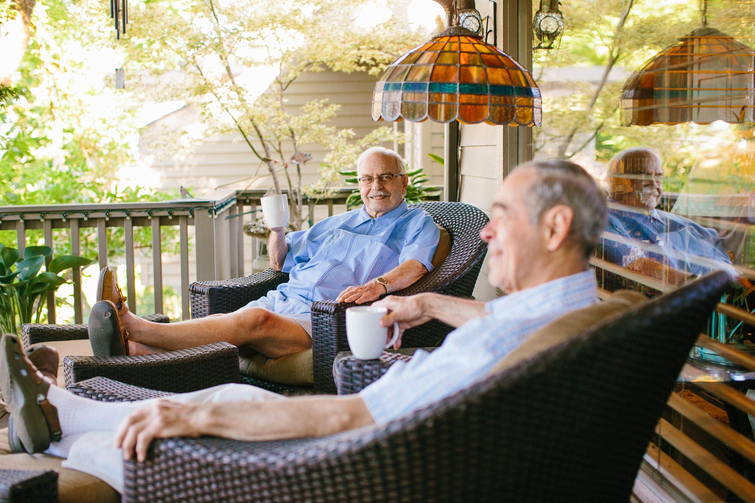 Two men sit and chat on the porch with coffee cups.