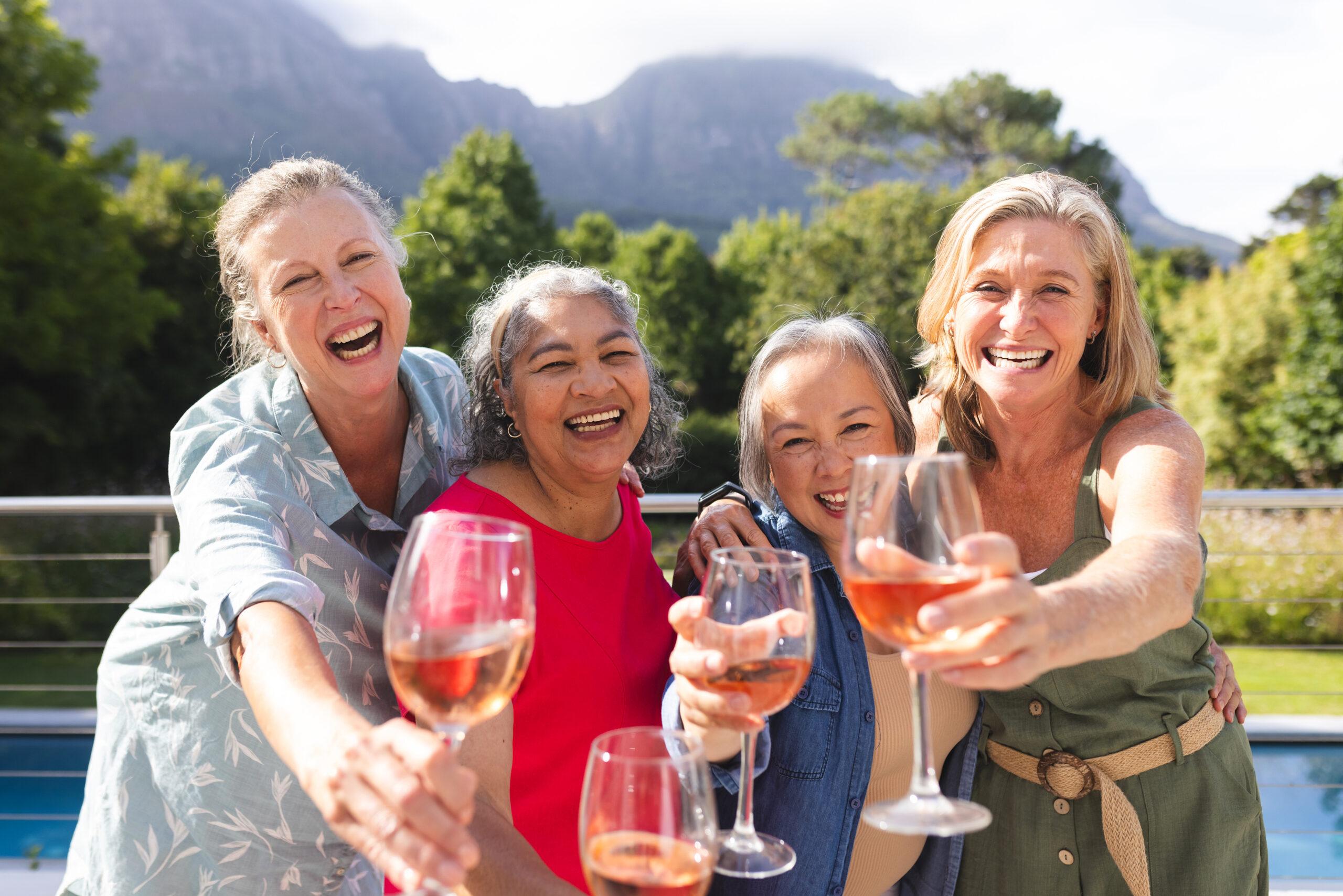 A group of four women smile and hold out wine glasses outdoors.