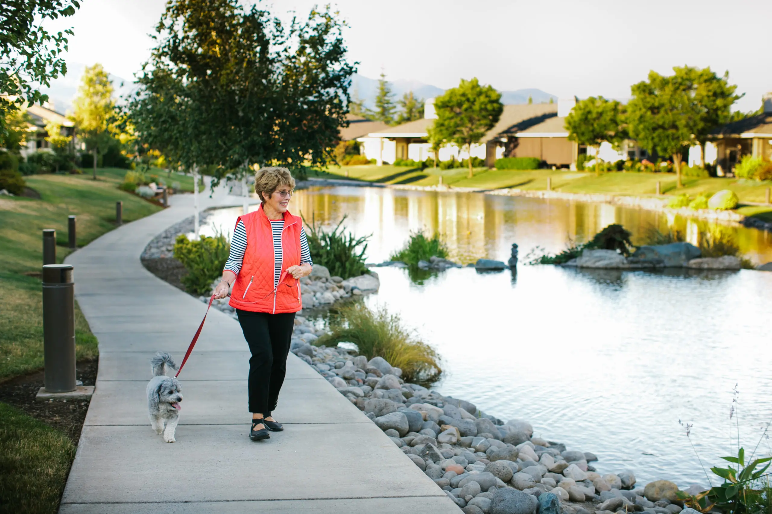 A woman wearing a coral colored vest walks a dog next to a picturesque lake.