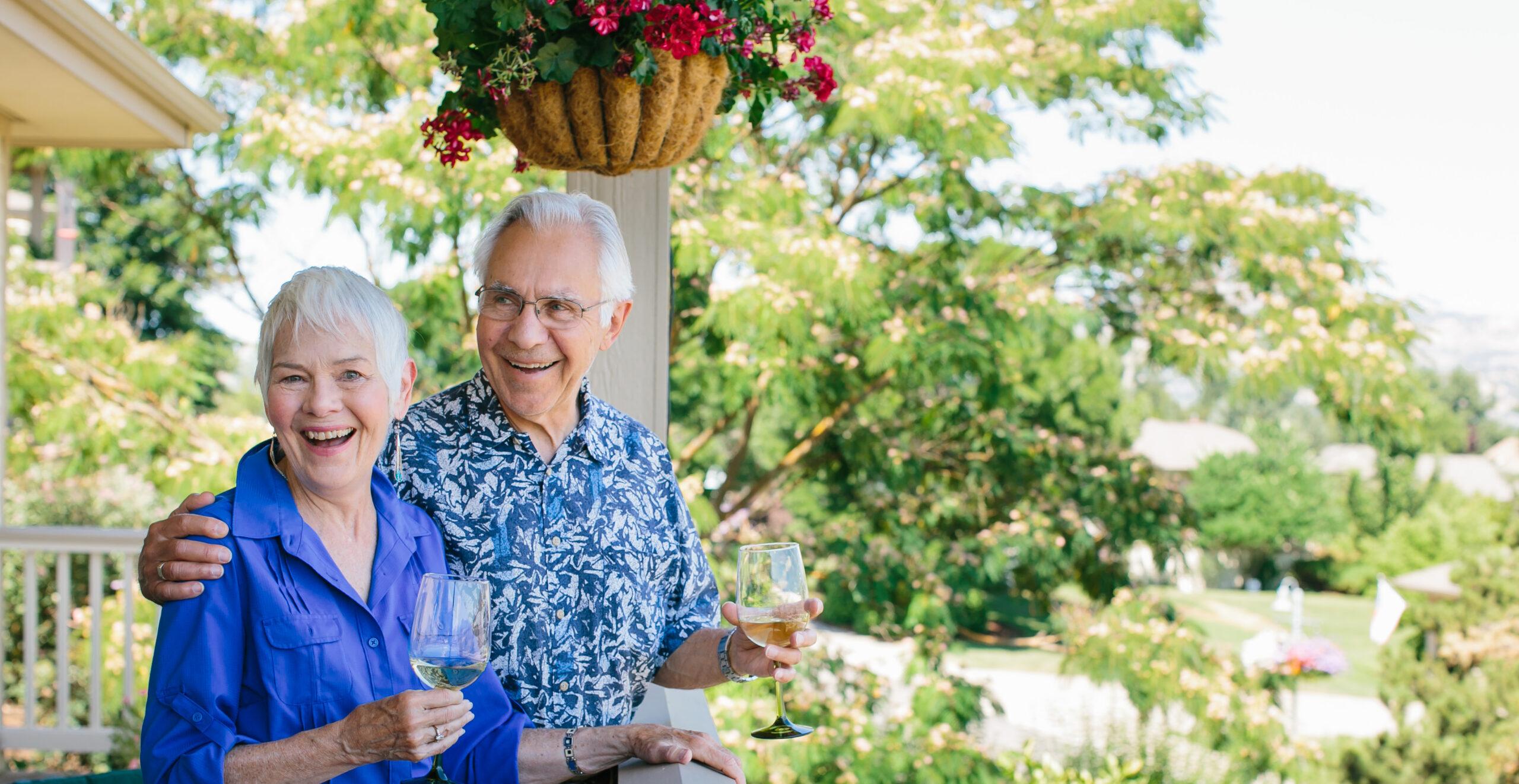 An older couple holding wine glasses smiles on an outdoor patio.