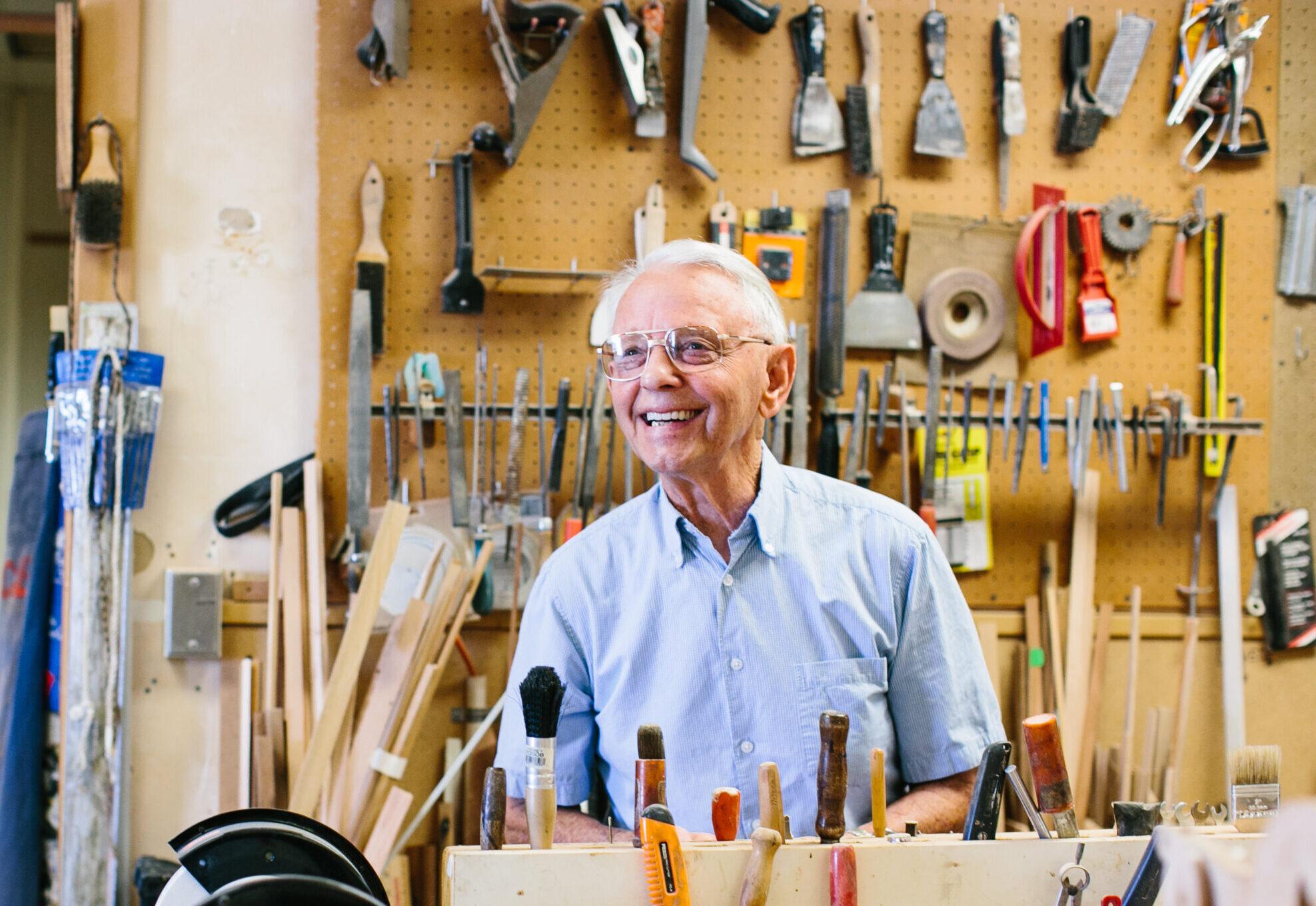 A smiling man works in a woodshop, surrounded by tools.