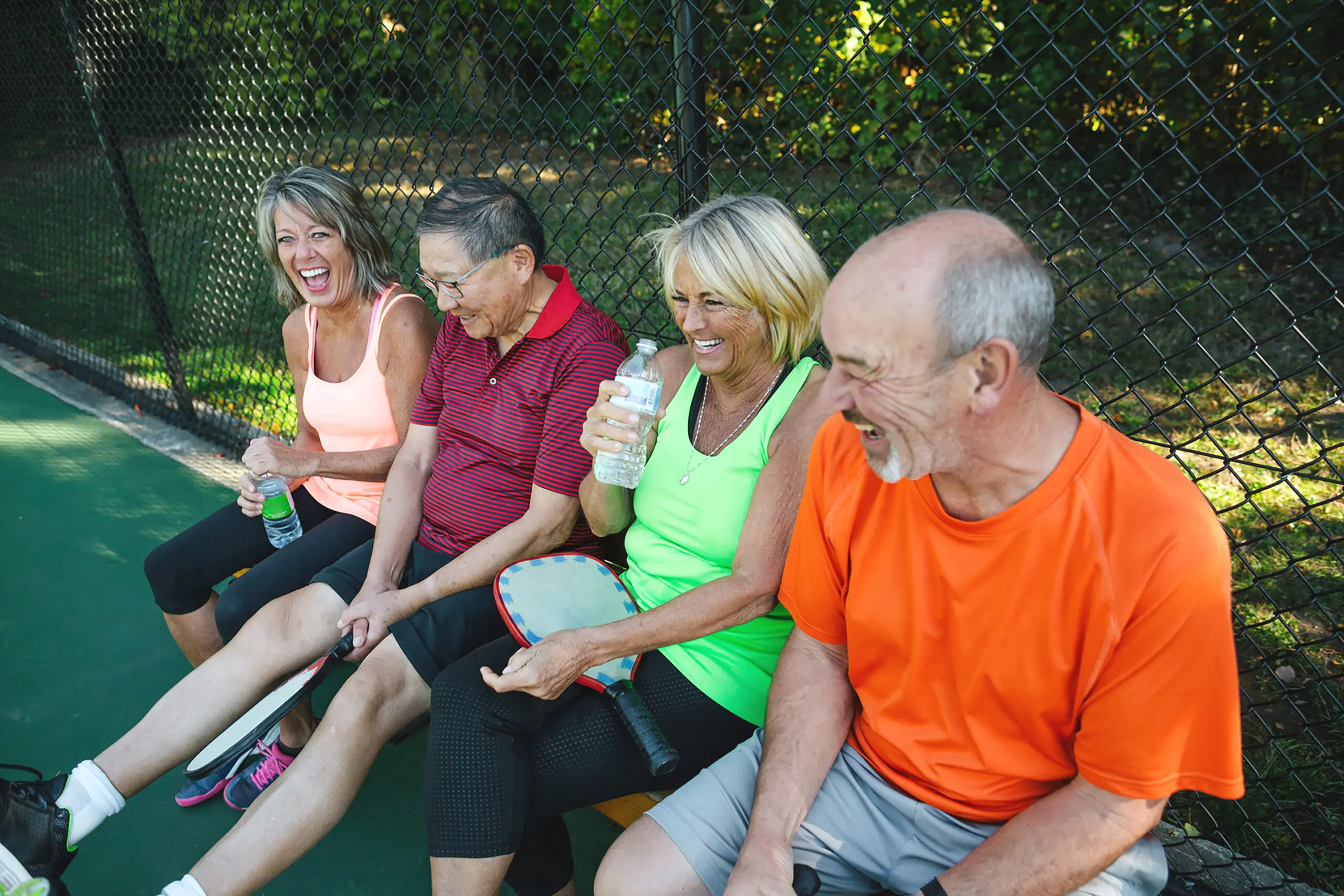 Four friends laugh and talk on a bench after a game of pickleball.