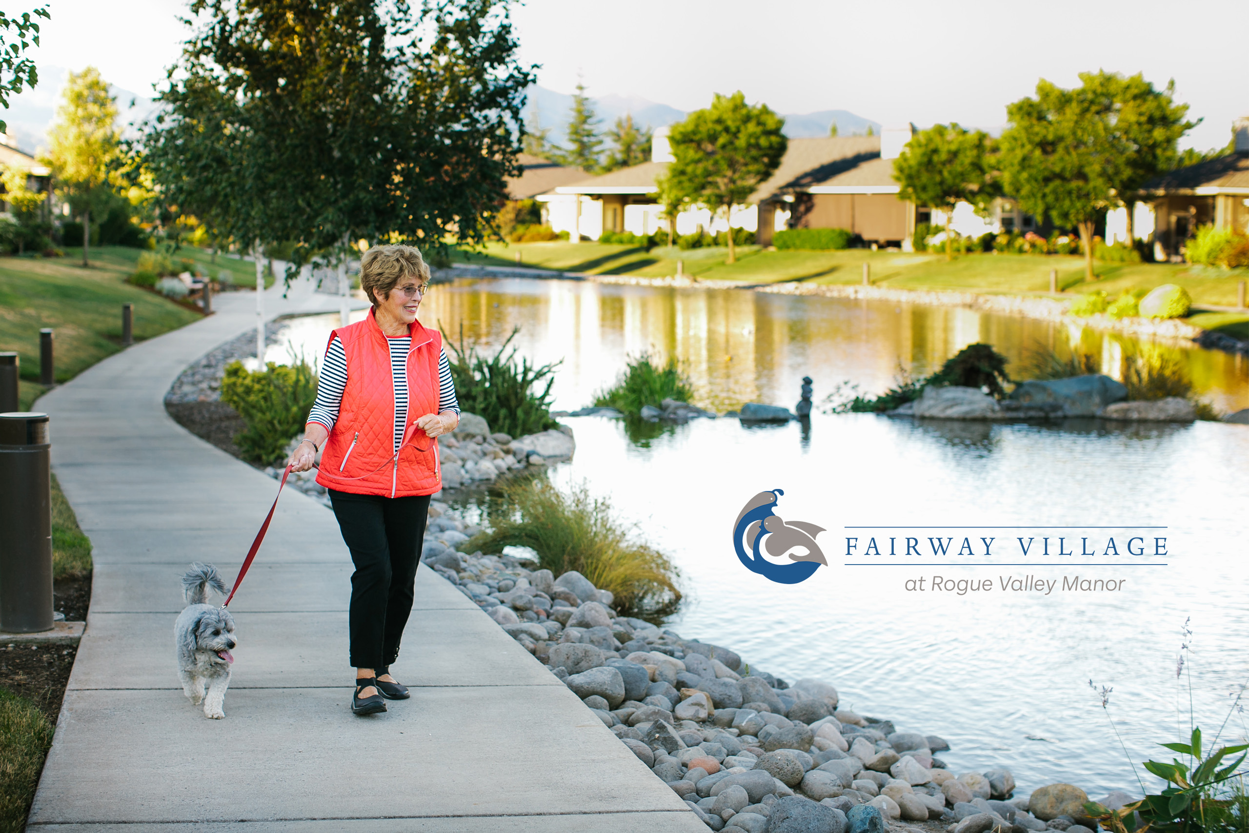 A woman wearing a coral colored vest walks a dog next to a picturesque lake.